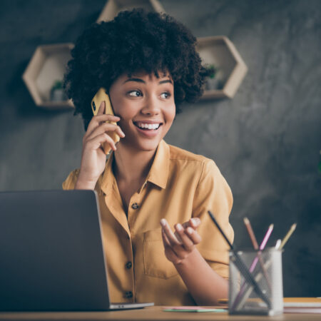 Photo of cheerful positive mixed-race pretty woman working as content manager, arranging with her customers by phone smiling toothily
